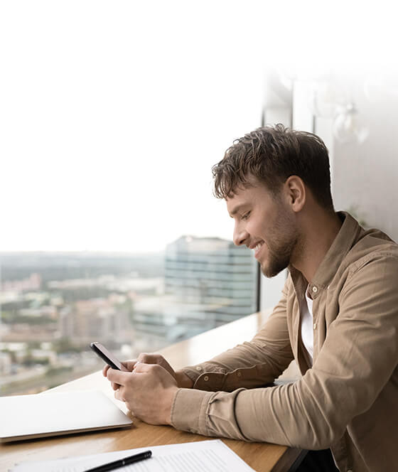 Een man zit in een café bij een raam, glimlachend en kijkend naar zijn smartphone.