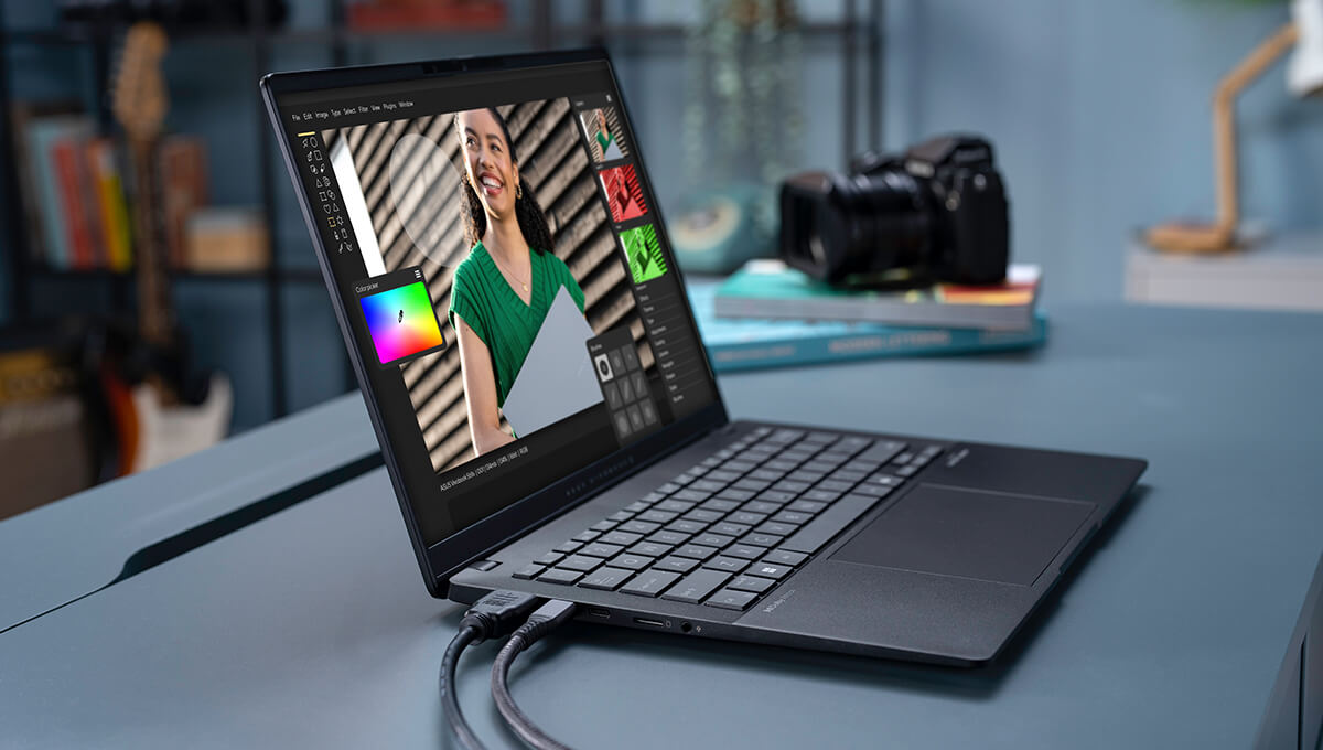 an ASUS Vivobook S 14 in neutral black on a desk, running an image editing application, with connected cables and a camera in the background.