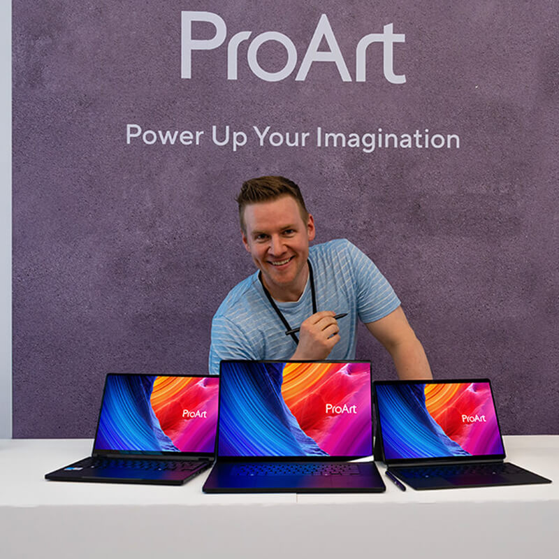 Jazza smiles holding a stylus behind three ProArt laptops.