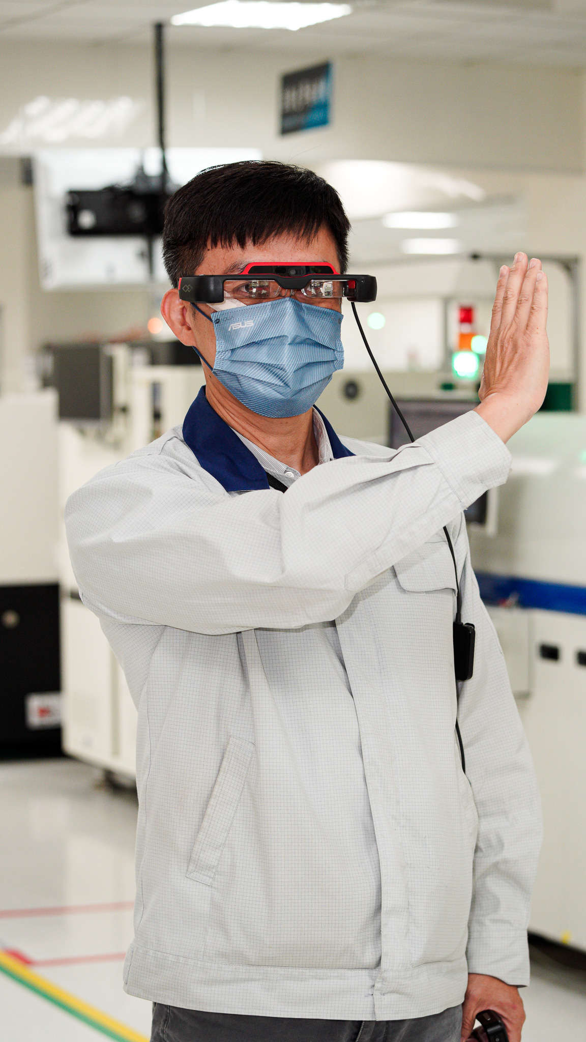 a worker in a factory wearing AR smart glasses and a blue surgical mask. The individual is engaged in an interaction machine using his hand