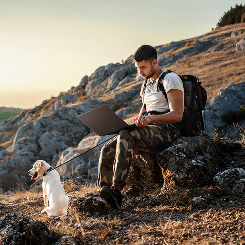 A man is sitting on a rock by a hiking trail with a ProArt laptop on his lap. A white Labrador is sitting in front of him looking out at the mountain scenery.