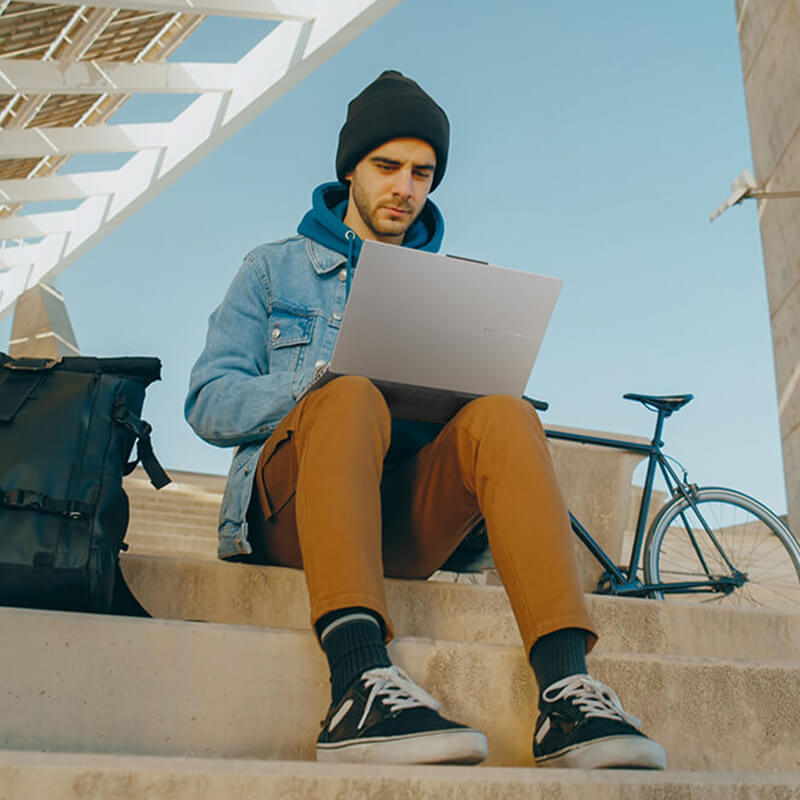 Young man sits on stairs outdoors with an ASUS Vivobook Pro 15 OLED laptop on his lap