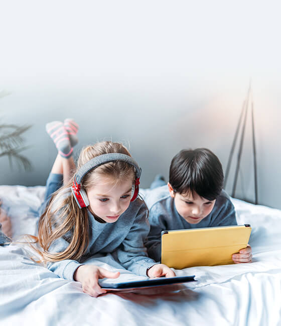 Two children lying on a bed using their tablets.