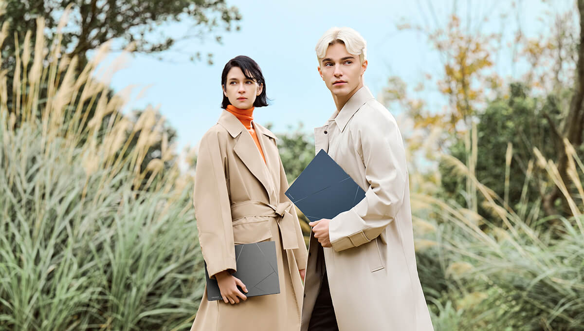 A man and a woman stand outdoors in a park-like setting, each holding an ASUS Zenbook.