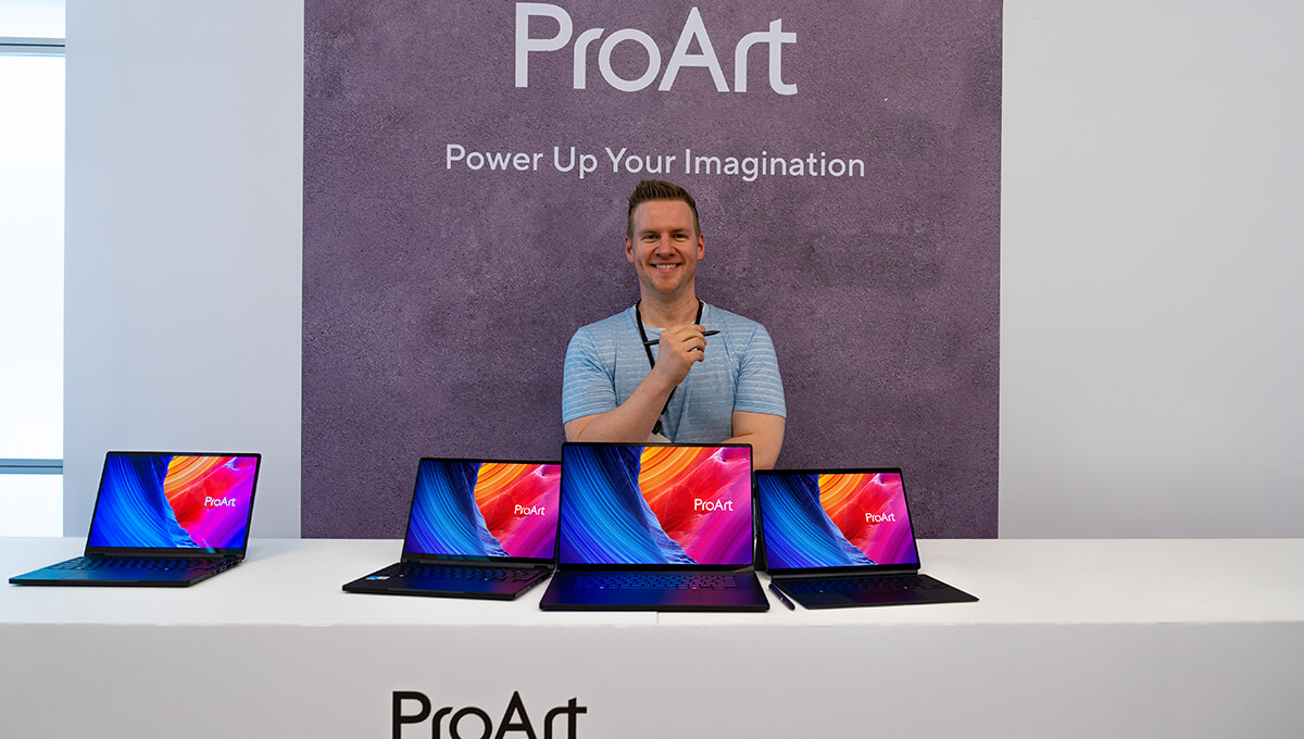 Jazza stands behind three ProArt laptops and smiles while holding an ASUS Pen 2.0.