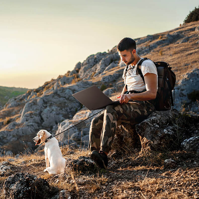 A man is sitting on a rock by a hiking trail with a ProArt laptop on his lap. A white Labrador is sitting in front of him looking out at the mountain scenery.