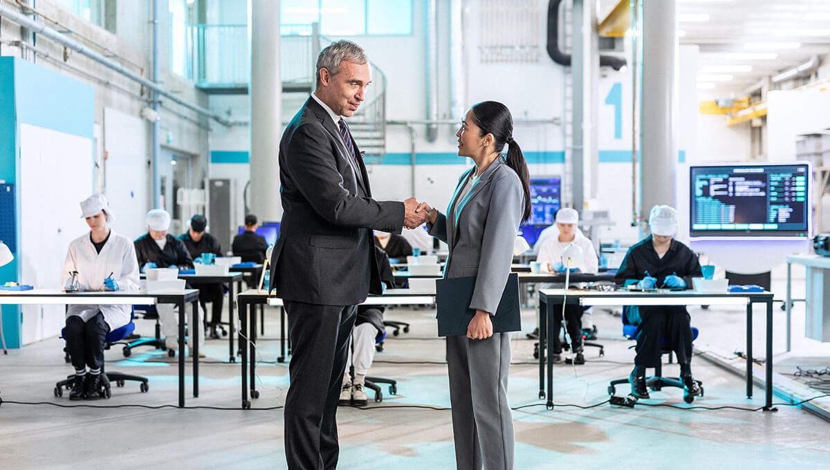 A man and a woman in suits are shaking hands in front of several rows of working people sitting at tables in lab coats.