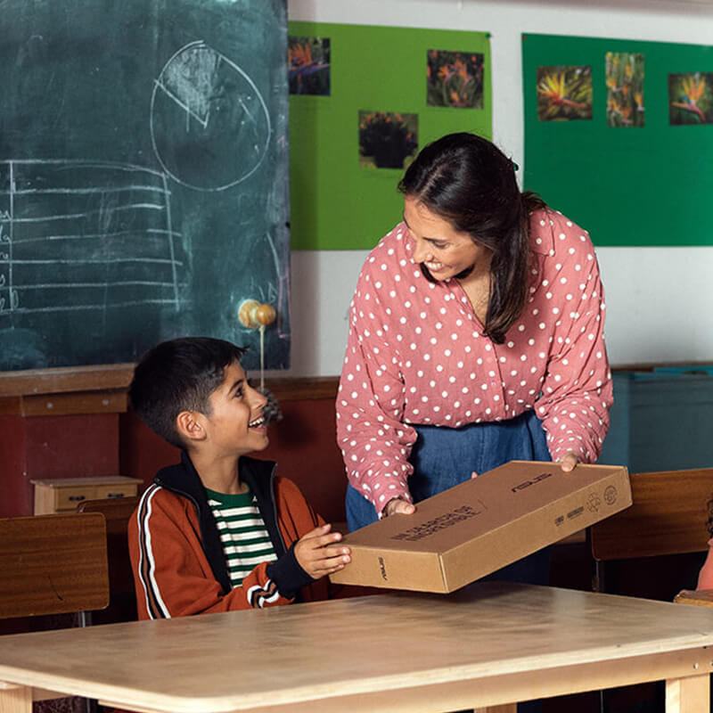 A female teacher in a classroom hands a young boy student a box that contains a laptop, and two young girl students smile as they use their ASUS laptops on a nearby desk.