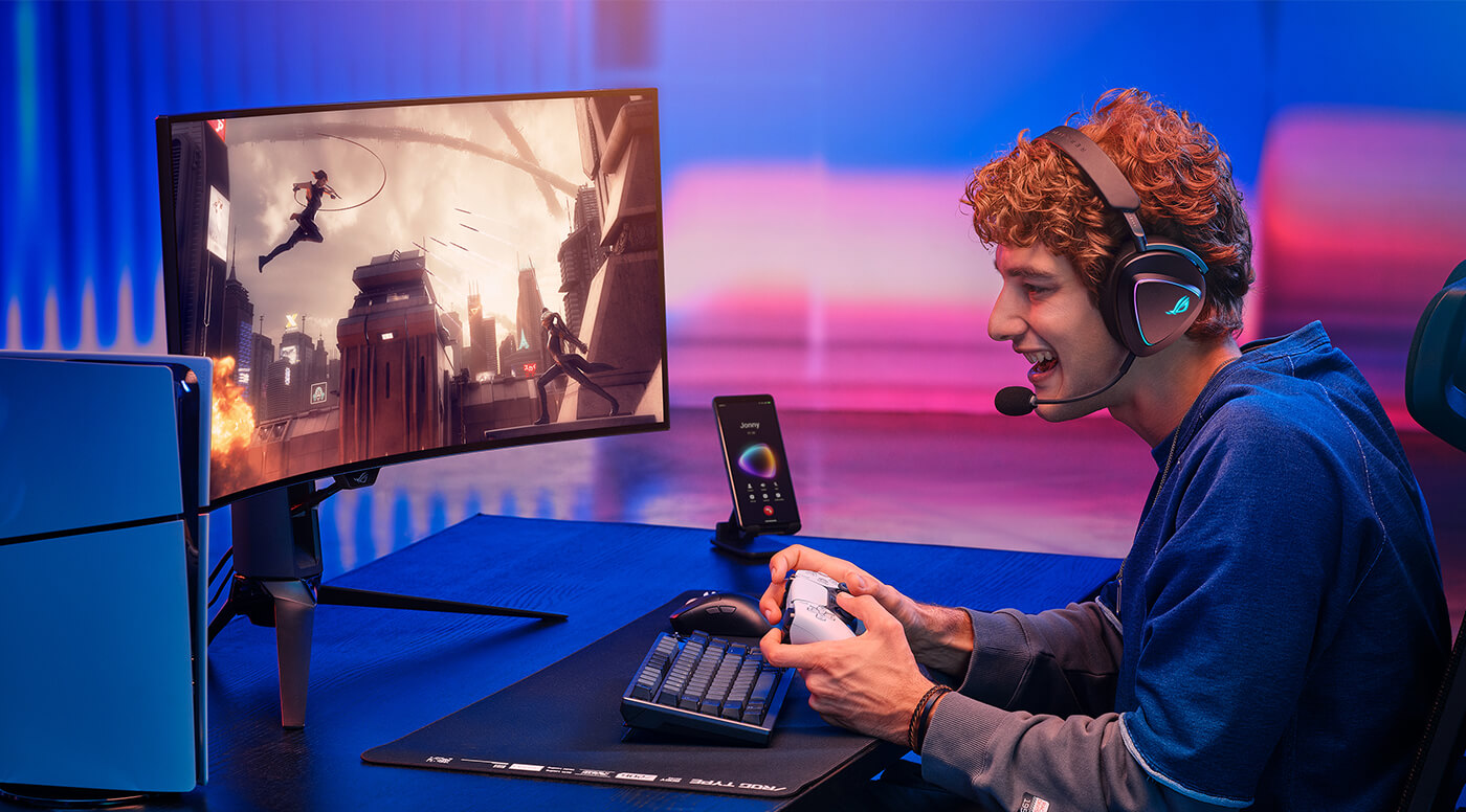 A man with curly hair wearing ROG Delta II holding a Playstation 5 controller, sits in from of the gaming desk with monitor beside to the Playstation 5. The ROG phone on the desk shows that man is in a phone call. The design mateirals on the top shows that he can swtich conversation to the Bluetooth mode when he picks up a phone call.