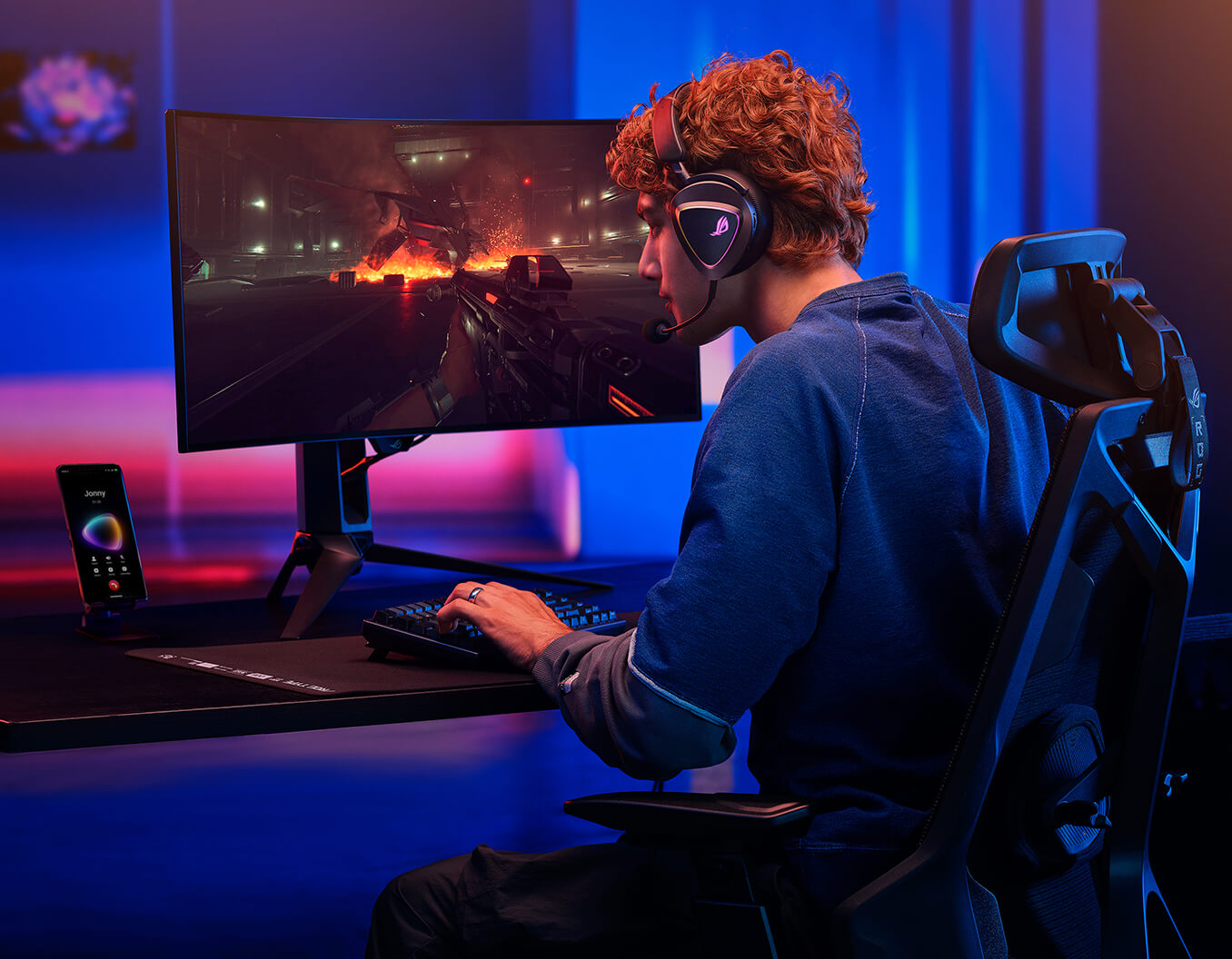 A man with curly hair sits at a ROG Destrier Core, wearing the ROG Delta II with a microphone and instense staring at the monitor. Viewing from the back, the ROG phone displaying the screen that he’s during a phone call.