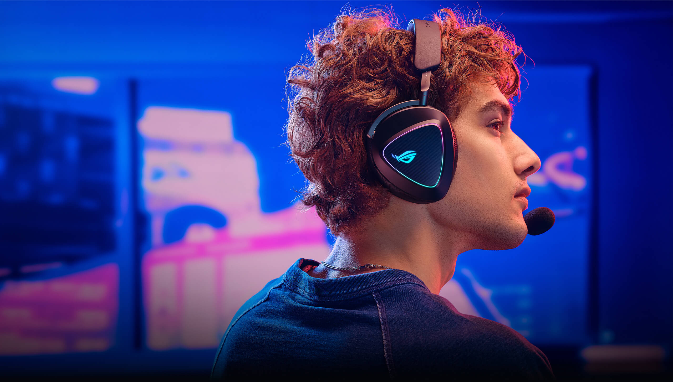 A man with curly hair wearing the ROG Delta II with the RGB lighting ROG logo on the ear cup, facing to the right. The background features a blurred neon gaming setup with blue and purple hues.