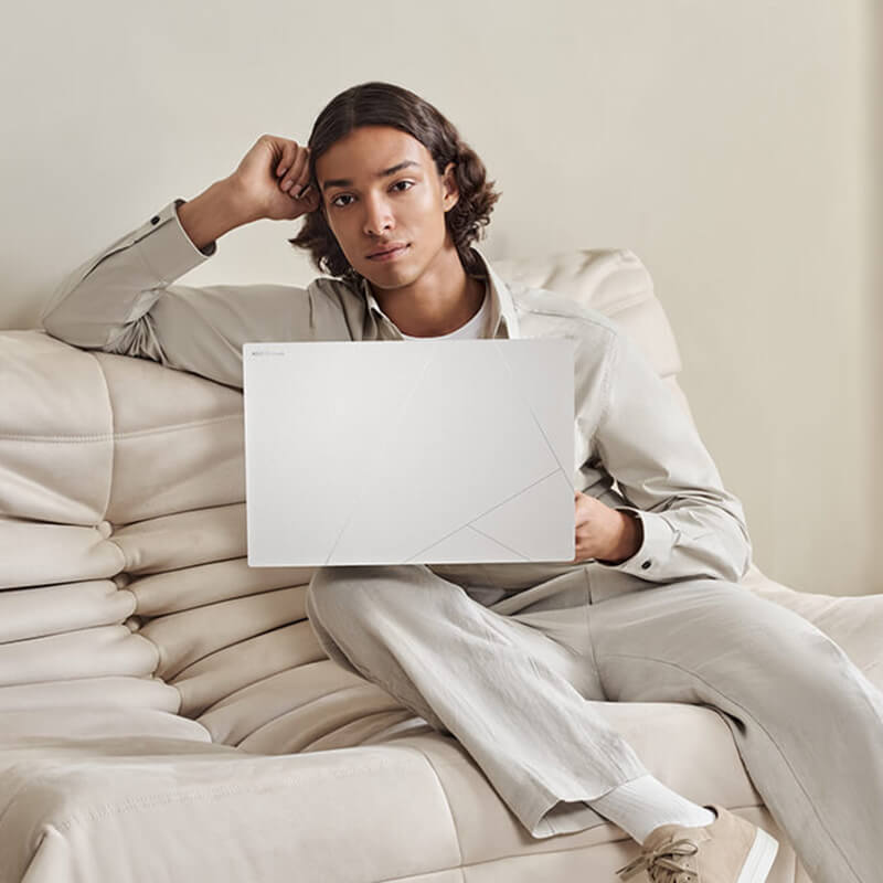 a young man sitting on a white sofa with a Zenbook S 16 laptop in Scandinavian White color on his lap