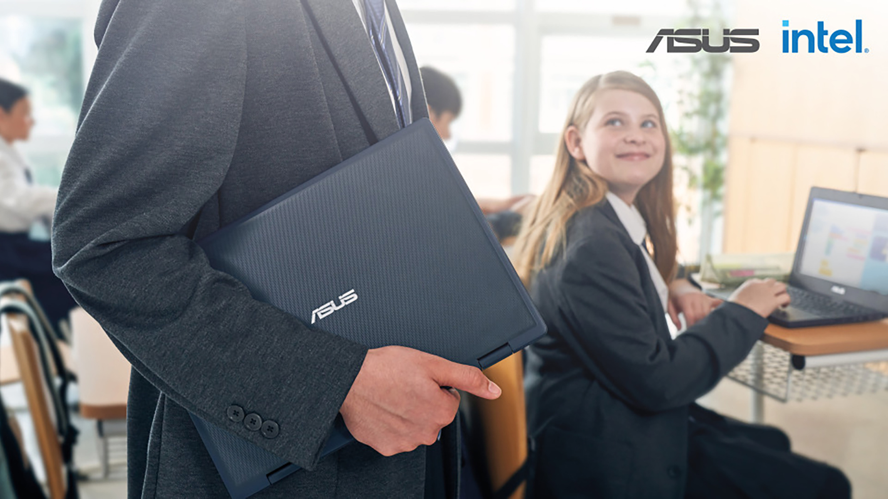 A teacher in a classroom holding an ASUS ruggedised laptop under his arm