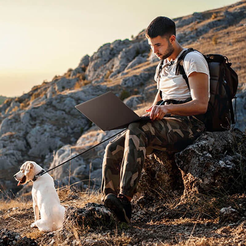 A man is sitting on a rock by a hiking trail with a ProArt laptop on his lap. A white Labrador is sitting in front of him looking out at the mountain scenery.