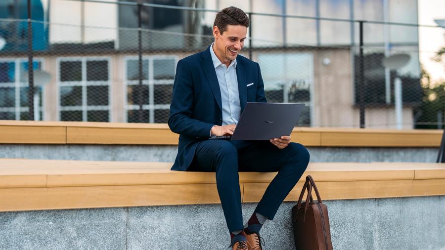 A man in the navy suit sitting on a long bench outdoor using the ASUS ExpertBook B9 on his lap, with a smile on his face.